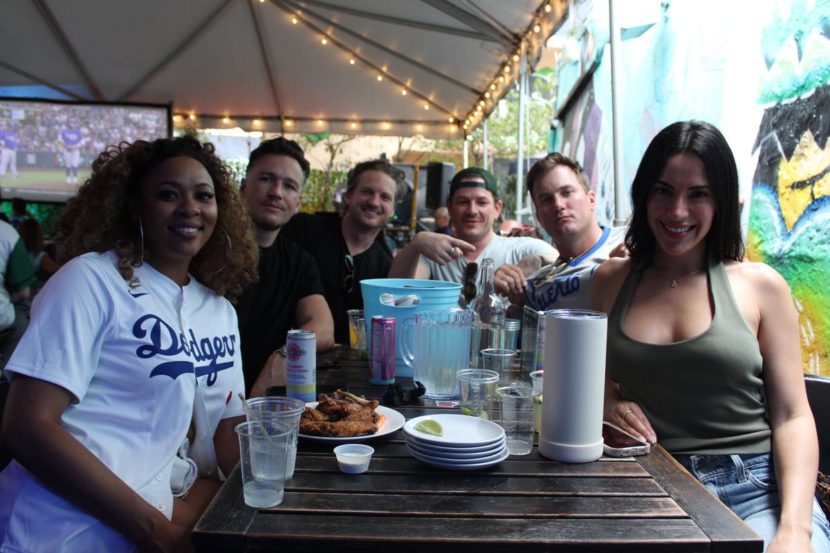 Group of friends in Dodgers and Puerto Rico jerseys around a Grails Wynwood patio table with wings, drinks and the MLB game on TV during a World Series watch party