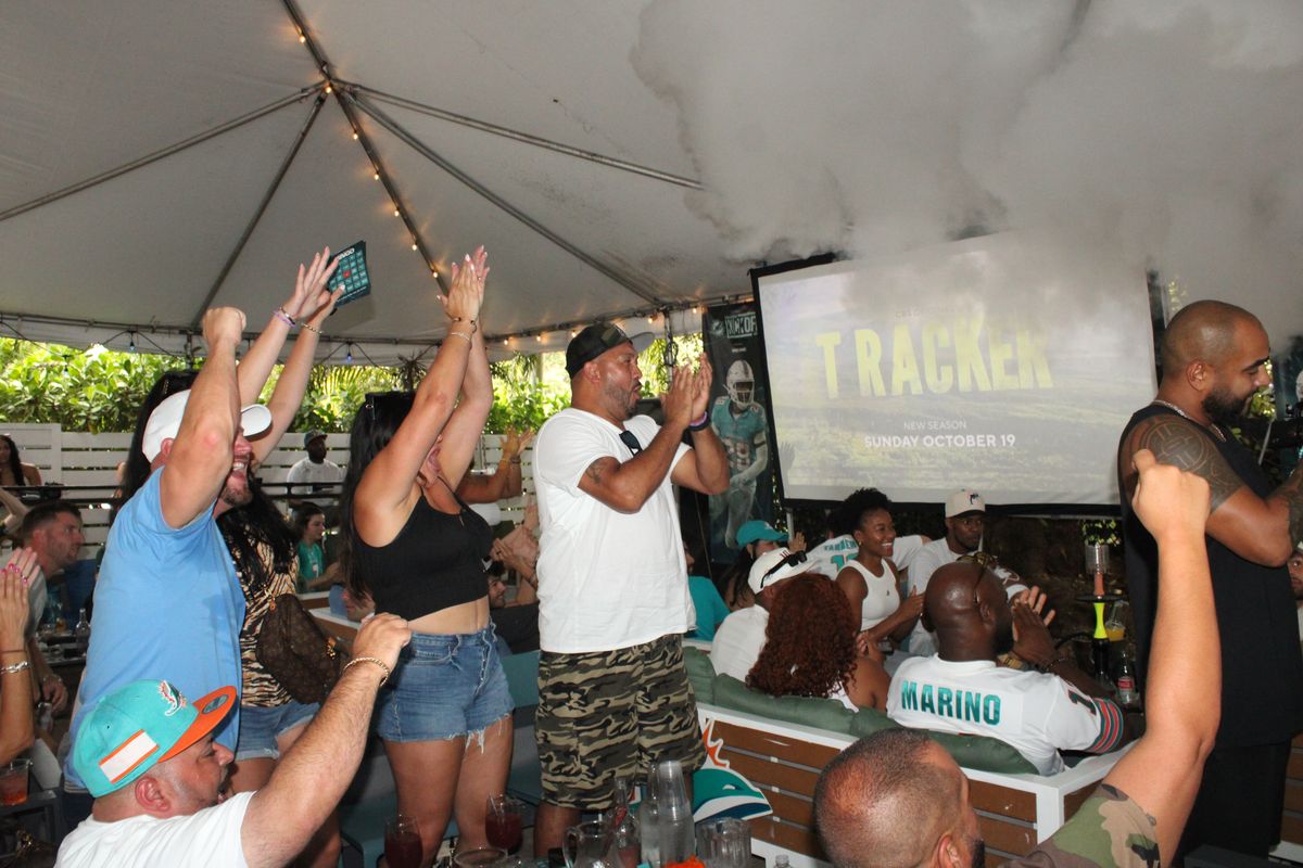 Miami Dolphins fans on their feet cheering with arms raised under the tented patio during a Dolphins watch party at Grails Miami