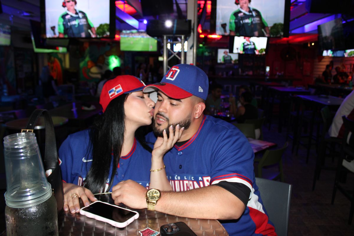 Couple in Dominican Republic baseball jerseys sharing a kiss at a Grails Wynwood table with the World Baseball Classic playing on the TVs