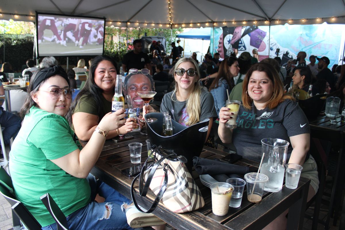 Four friends toasting drinks at a patio table, one in a Philadelphia Eagles tee, with the NFL game up on the giant projector screen behind them at Grails Miami