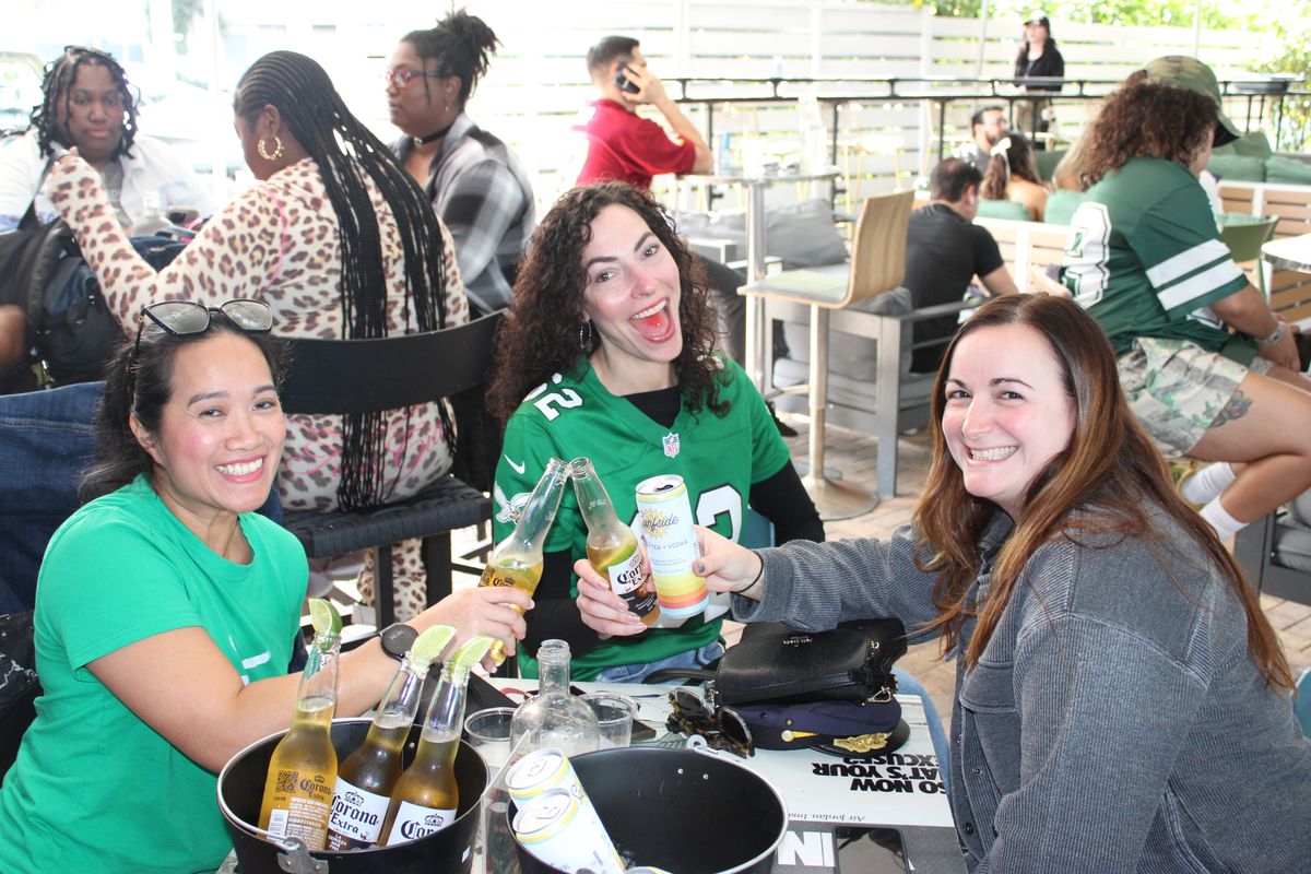 Three Philadelphia Eagles fans in green toasting cold beers at a patio table on an NFL Sunday in Miami