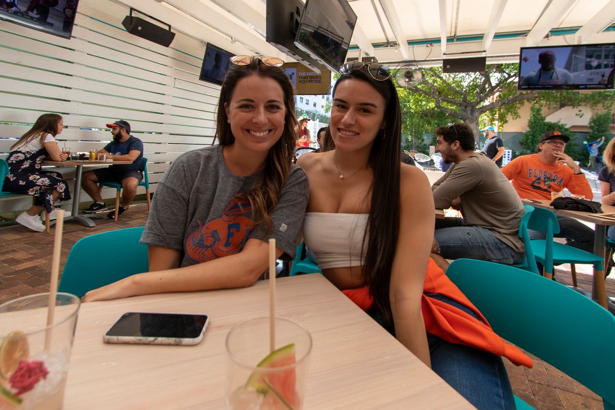 Two friends at a Grails Miami patio table, one in a grey Florida Gators shirt, with cocktails on a Gators game day