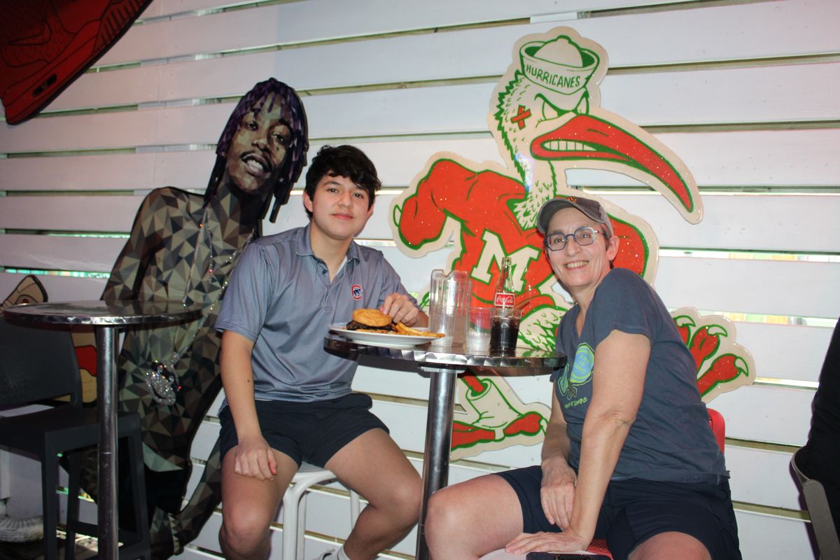 Mom and son eating burgers in front of the Miami Hurricanes mural at Grails Wynwood on a college football Saturday