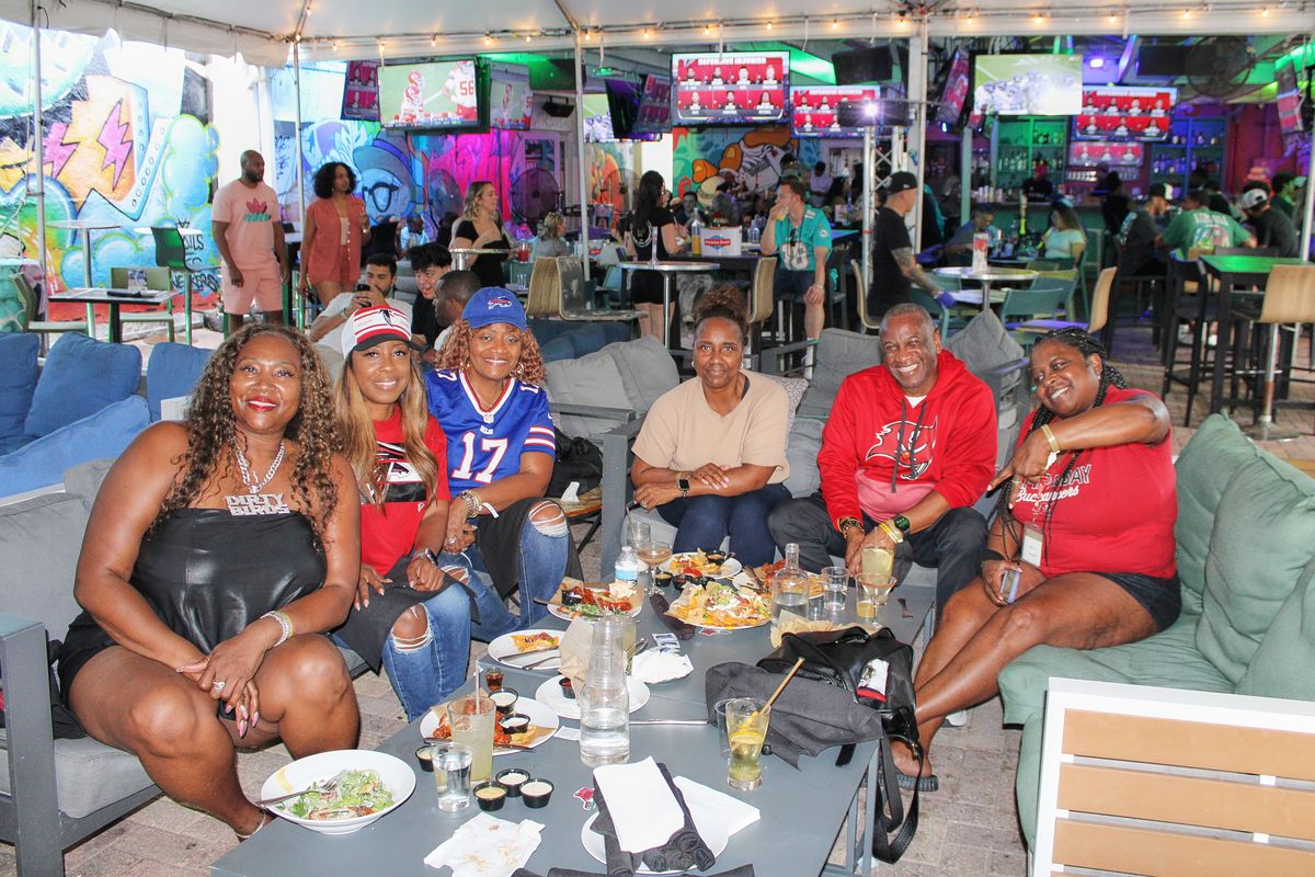 Group of six NFL fans, one in a Buffalo Bills number 17 jersey and Bills cap, around a shared table under the tented Yard at Grails Miami