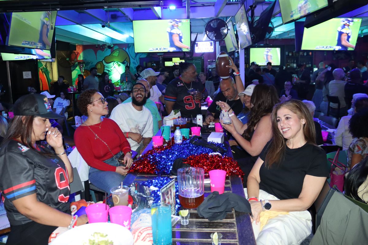 Friends in NFL jerseys laughing around a pom-pom covered table with football on every screen at the Thursday Night Football bar in Wynwood, Miami