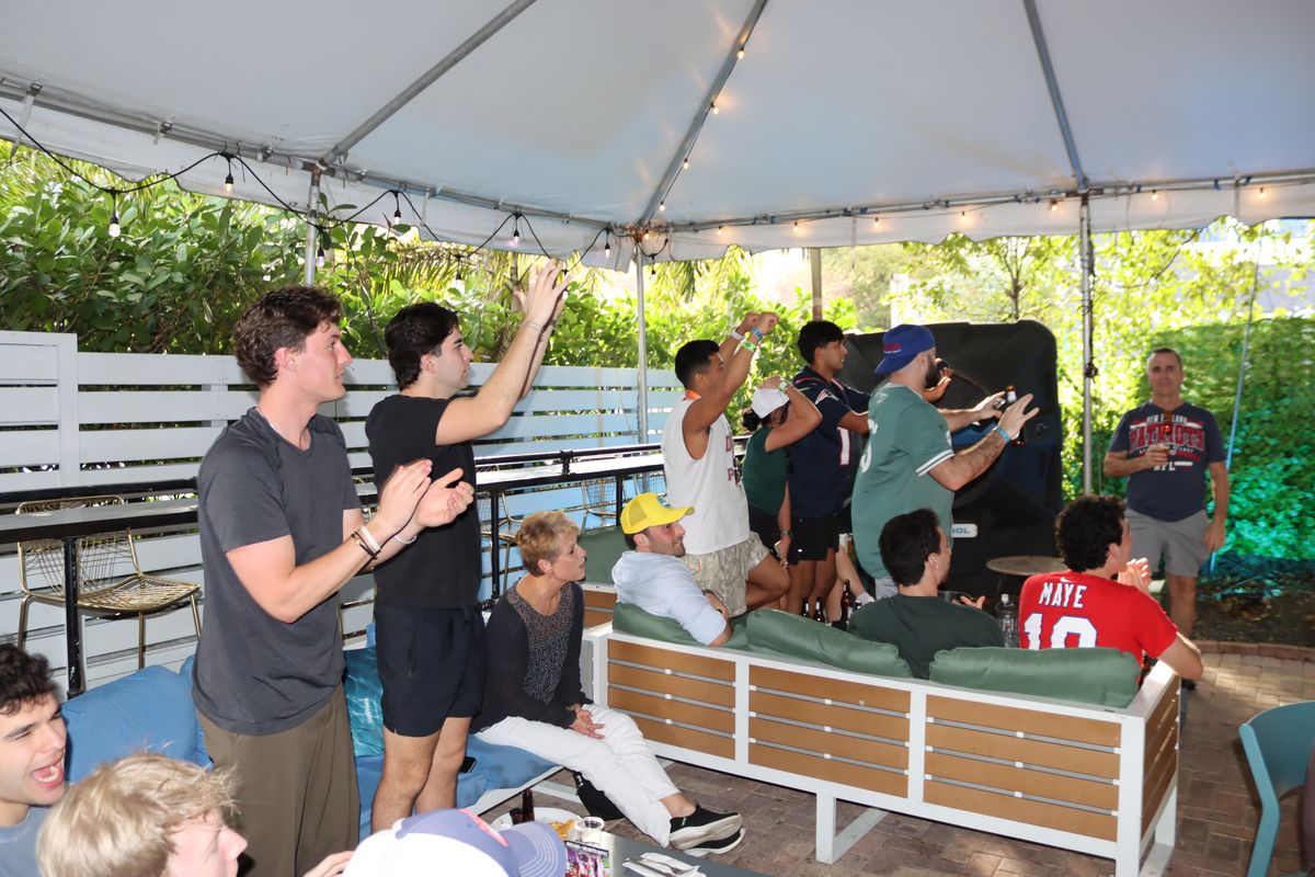 Football fans on their feet with arms raised celebrating a touchdown under the covered patio at Grails Miami