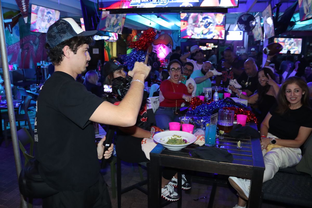 Watch party host with a microphone and pom-poms hyping a table of cheering football fans at Grails Miami