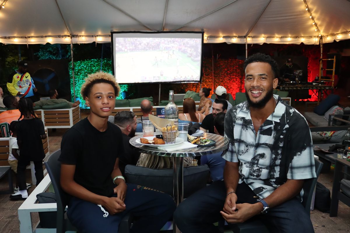 Two friends at a high-top table with the football broadcast on the big screen behind them at Grails Miami in Wynwood