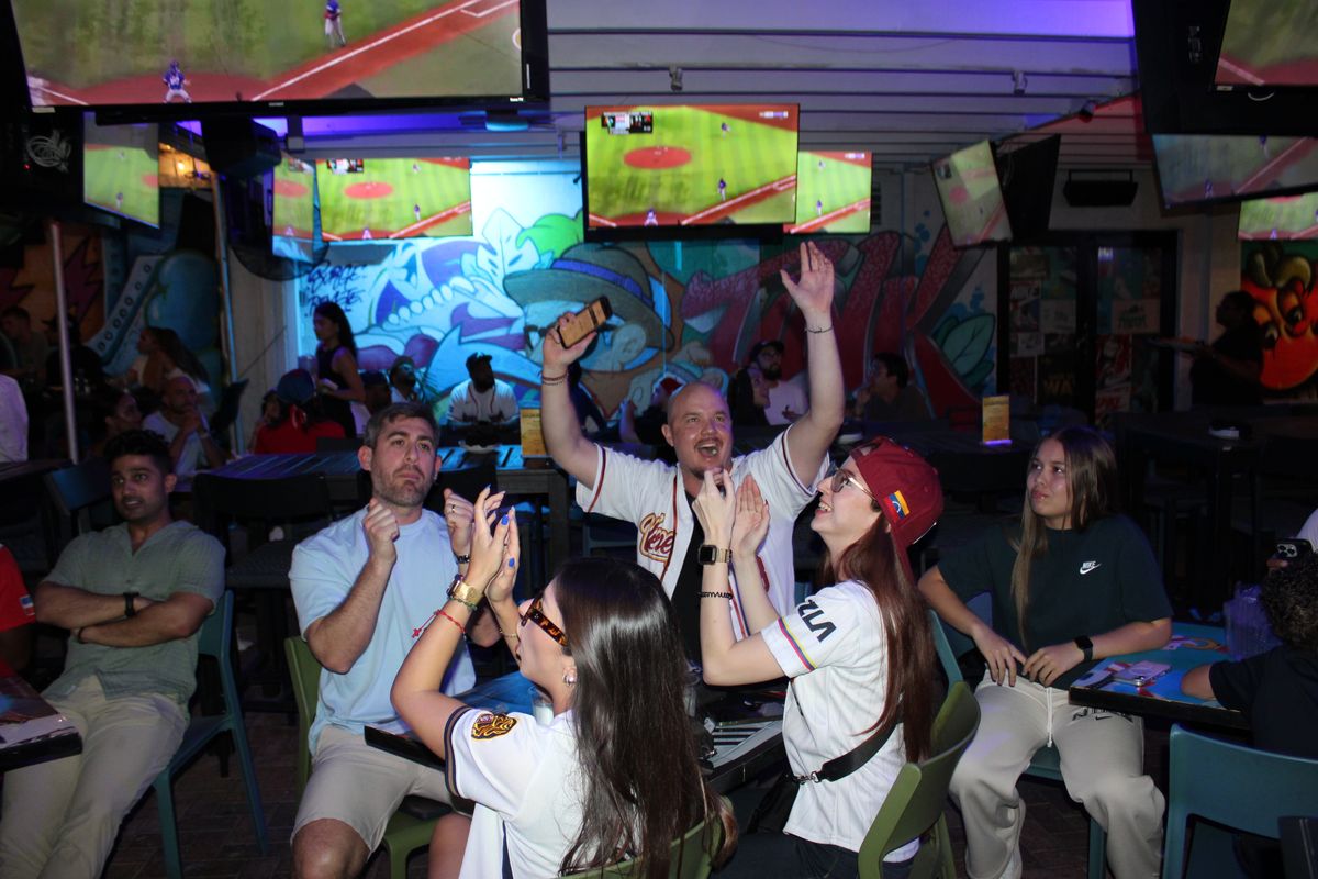 Venezuela baseball fans in team jerseys cheering and clapping while watching the game on multiple overhead TVs at Grails Wynwood during a Miami Marlins watch party