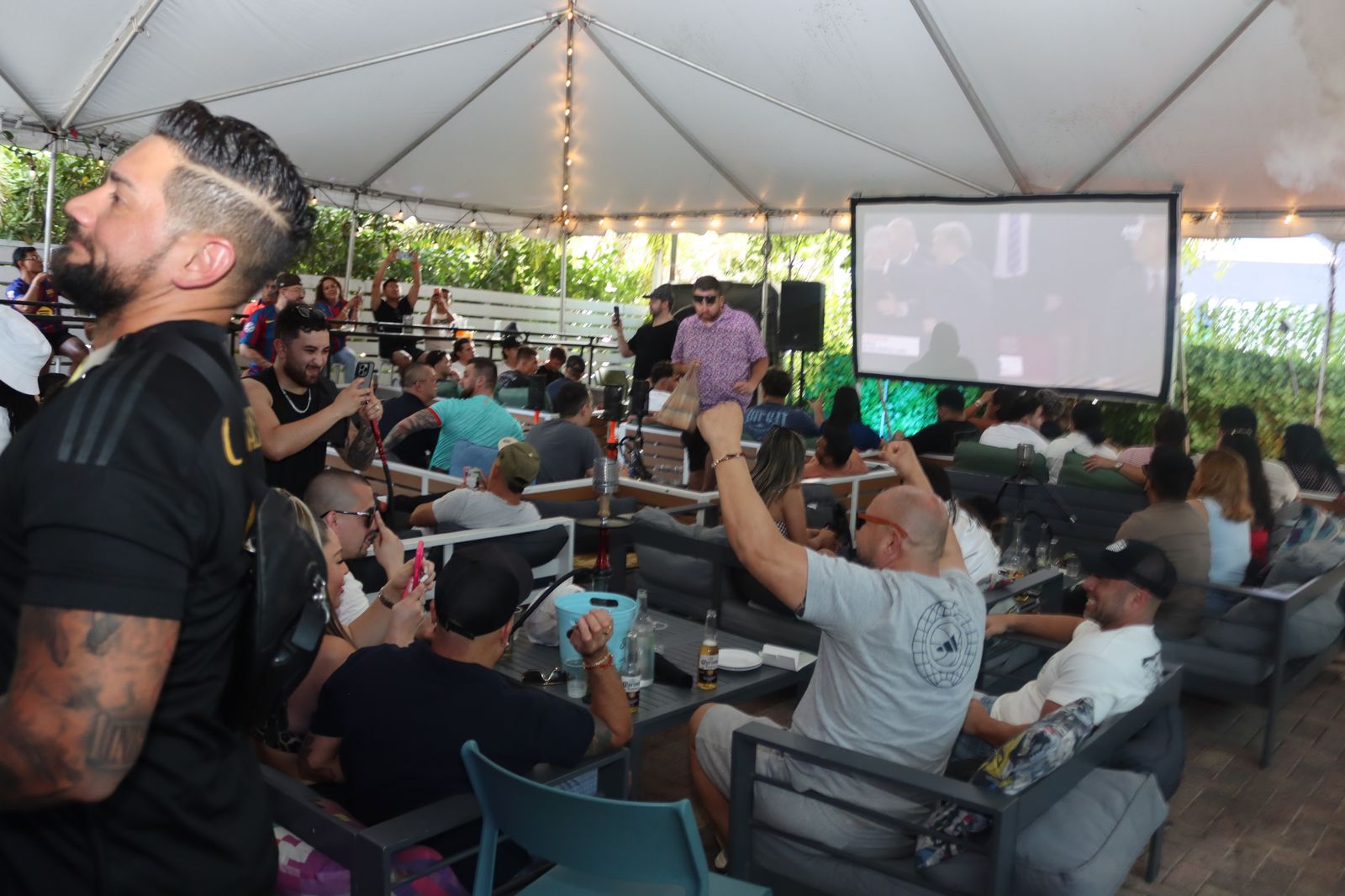 The Yard at Grails Miami Wynwood: the covered tent with the giant projector screen carrying a soccer match, a crowd of fans watching from sofa seating with hookah and string lights overhead.