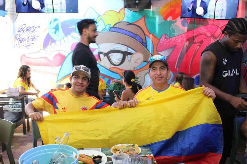Two Colombia fans holding up the Colombian flag at their table during a Colombia watch party in Wynwood, Miami