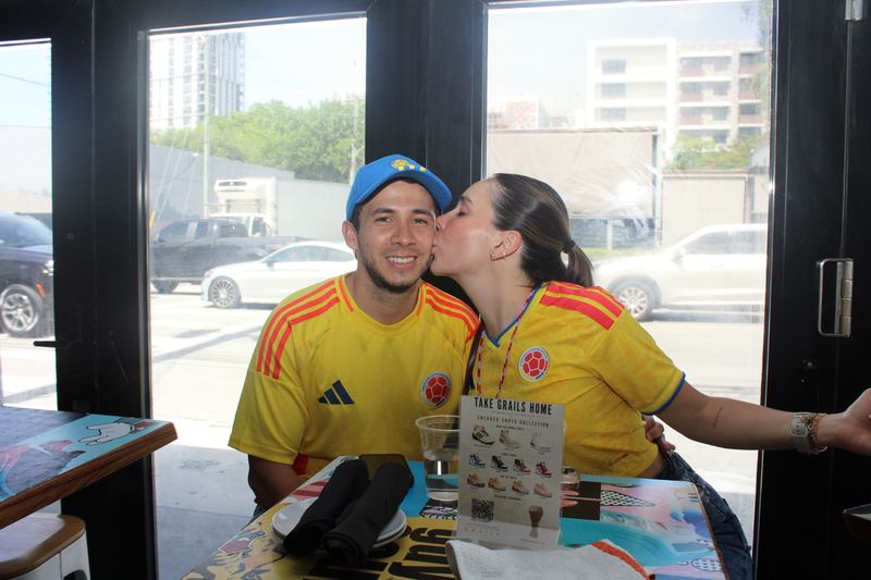 Couple in Colombia jerseys sharing a kiss at a window table during a Colombian soccer watch party at Grails Miami