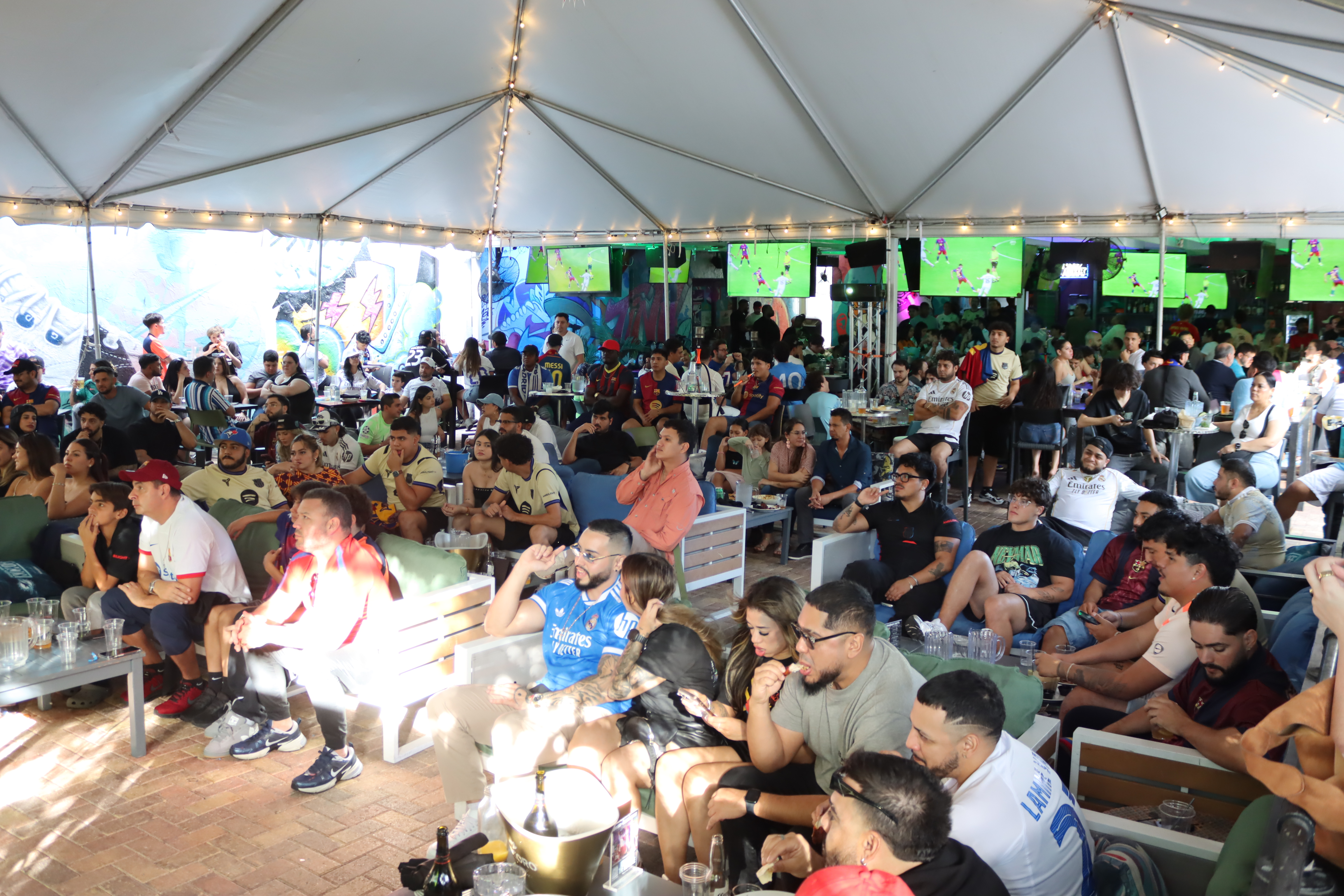 Packed crowd of soccer fans in Real Madrid and Barcelona jerseys watching El Clasico on multiple TVs under the tented patio at Grails Miami.