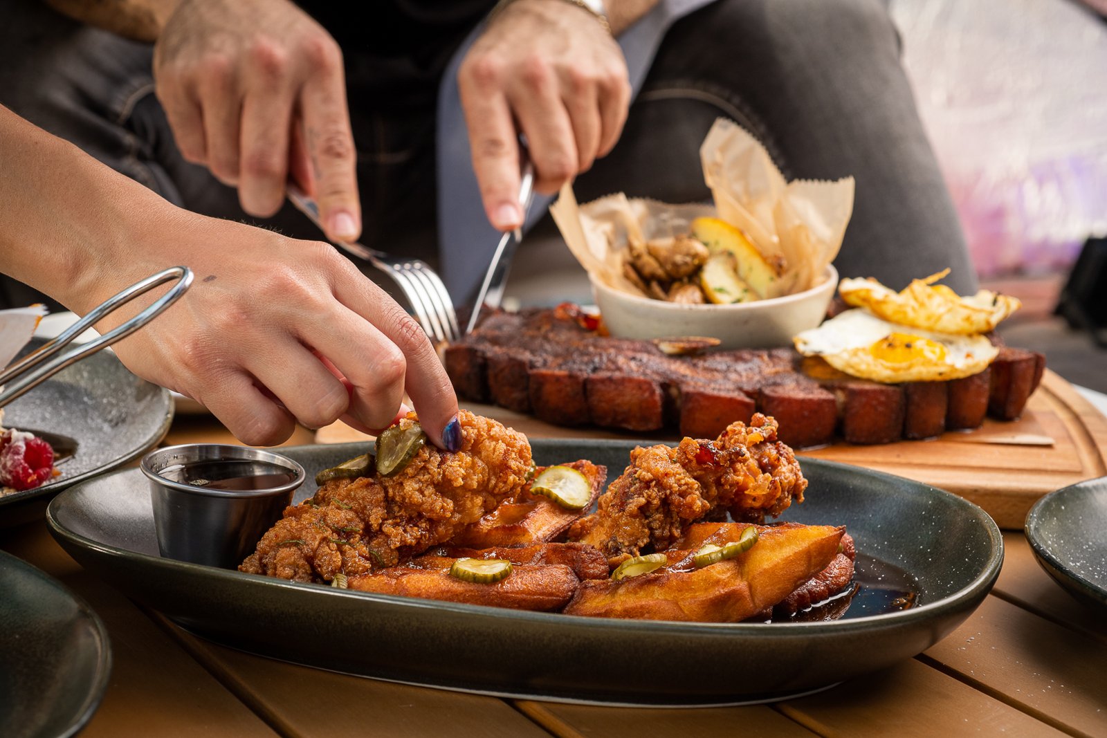 Woman reaches for a piece of fried chicken atop french toast with pickles and syrup while a second diner cuts into a steak with fried egg in the background.