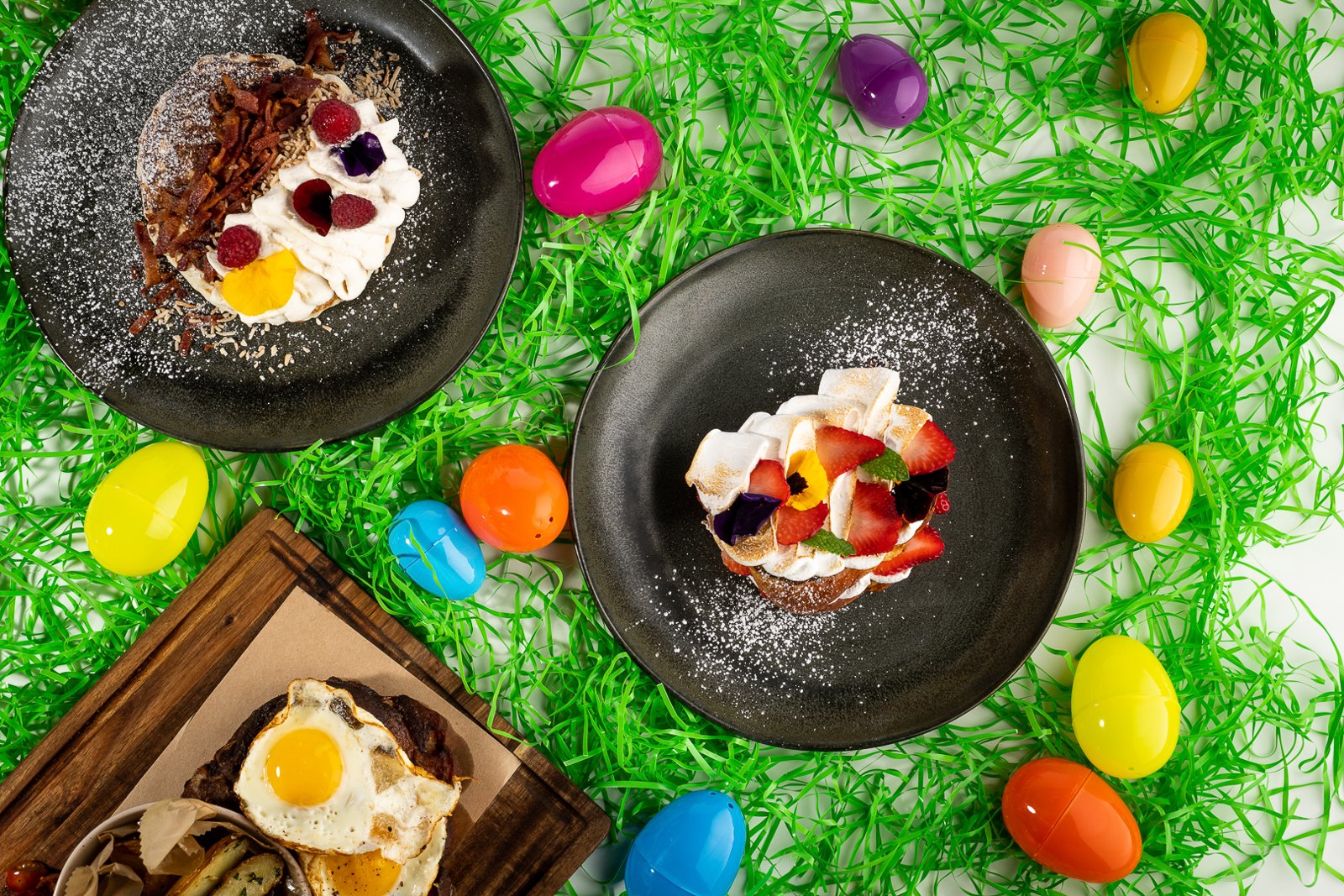 Overhead shot of Easter brunch plates with pancakes, pastries topped with berries, and a fried-egg dish surrounded by colorful plastic eggs on green grass