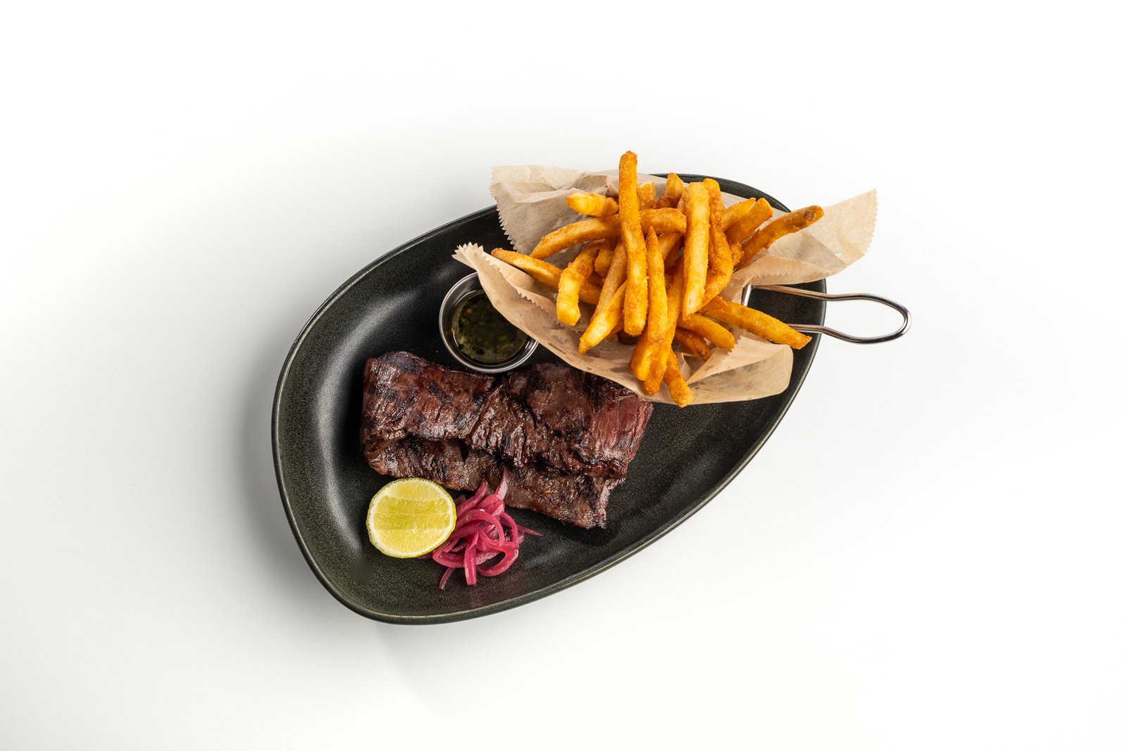 Grilled skirt steak with golden french fries, chimichurri sauce, lime and pickled onions on a black oval plate.