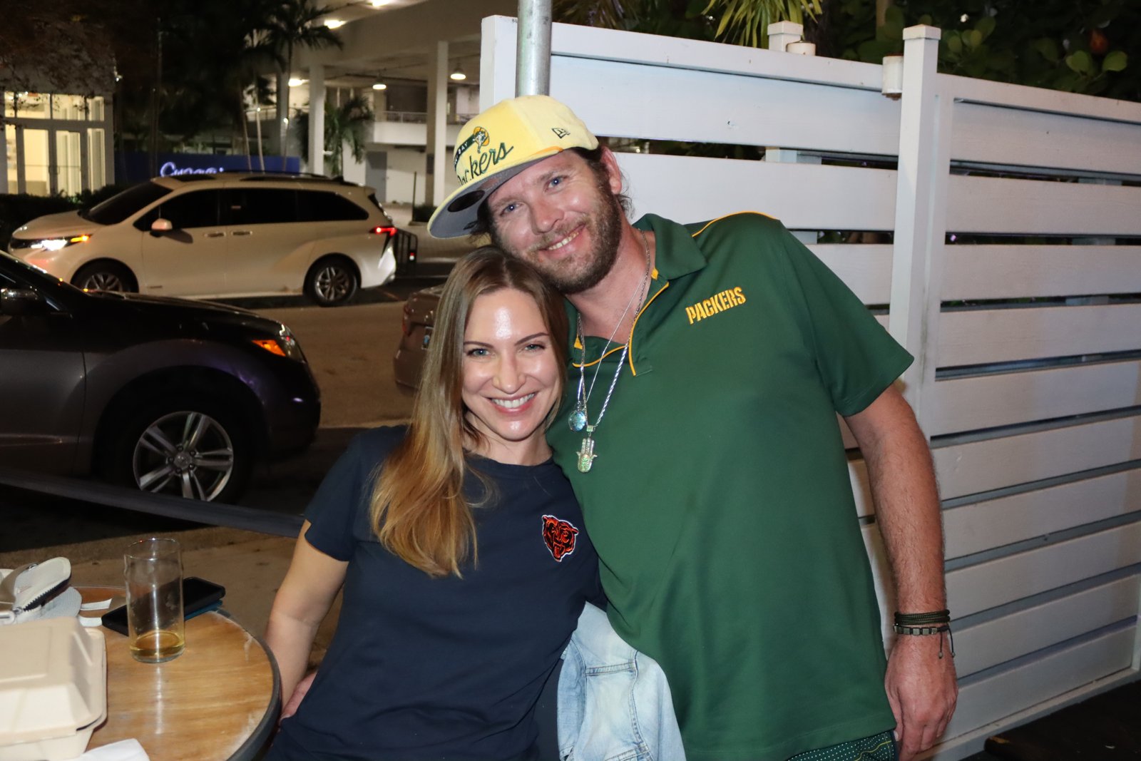 Smiling woman in Chicago Bears tee leans into man in Green Bay Packers polo and cap on Grails patio at night