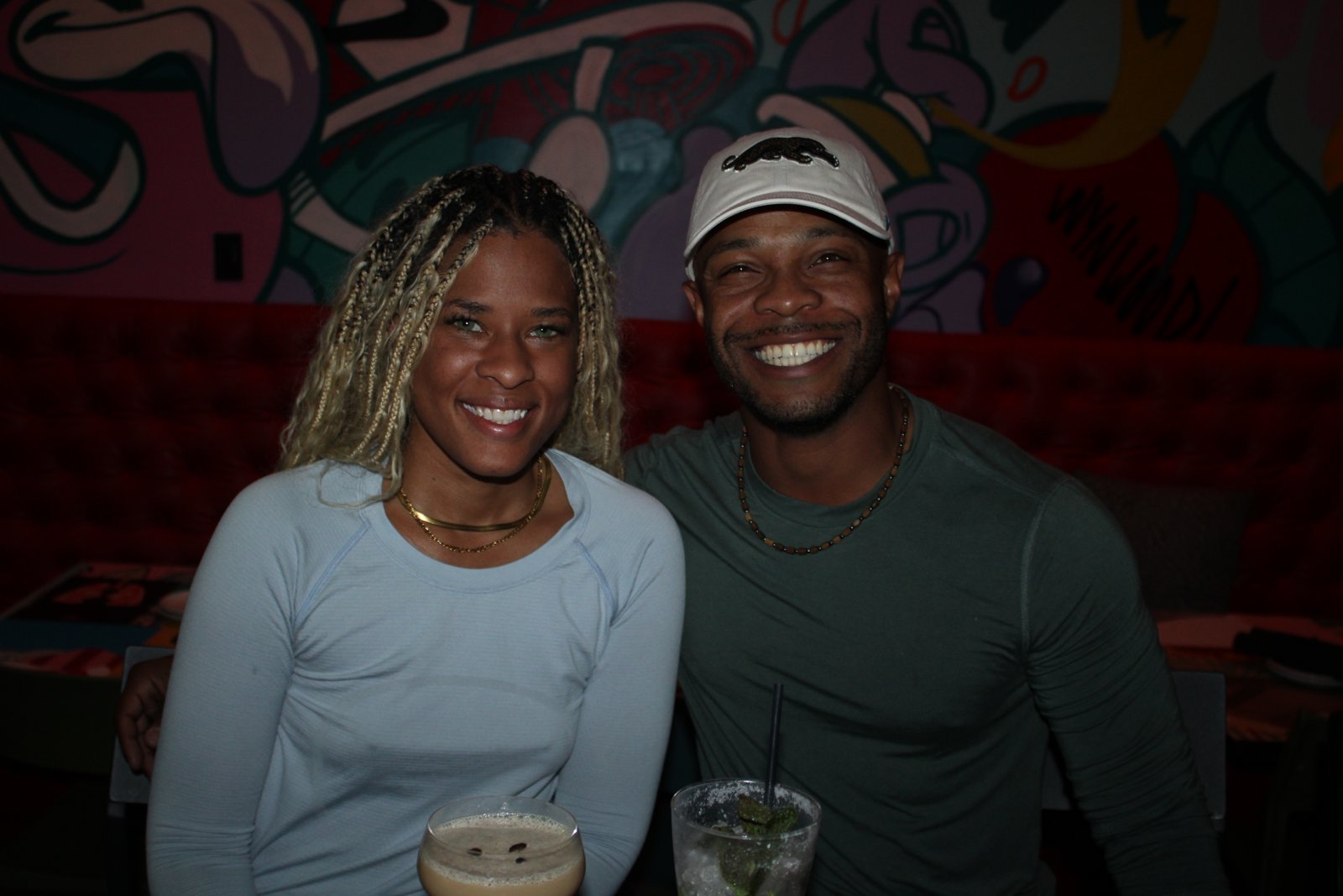 Smiling couple poses with an espresso martini and mojito at a red booth in front of a colorful Wynwood mural at Grails Miami.