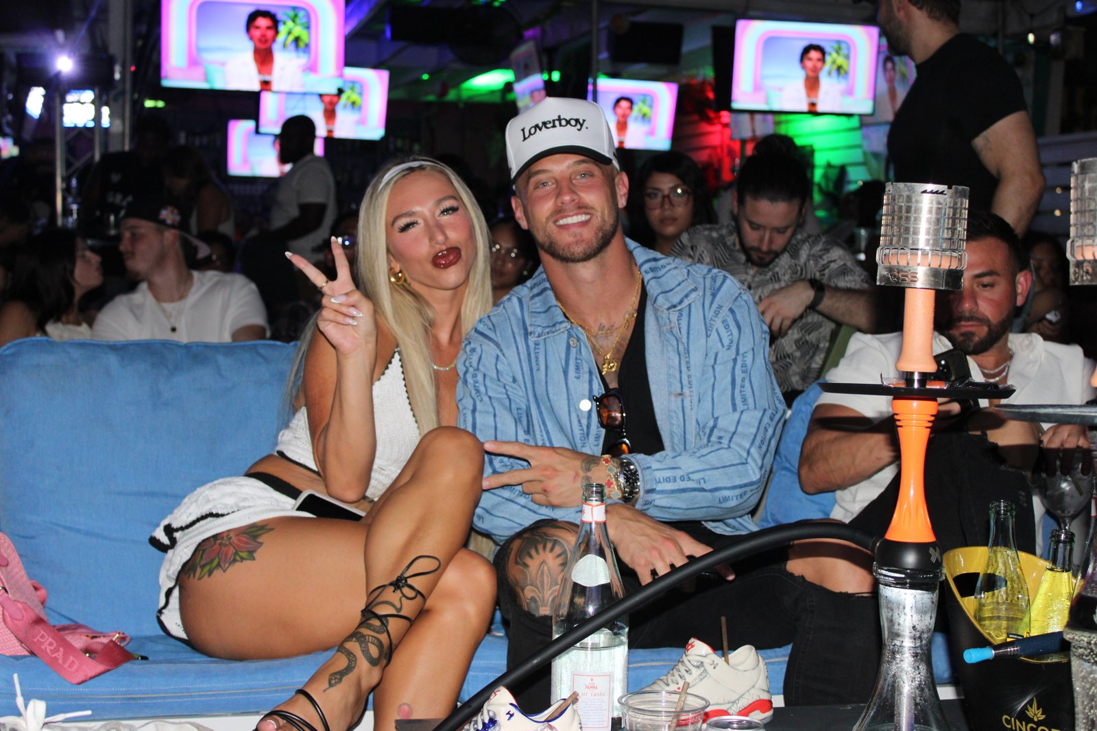 Blonde woman throws peace sign next to smiling man in Loverboy cap on a blue lounge couch with hookah and bottle service at Grails Miami.