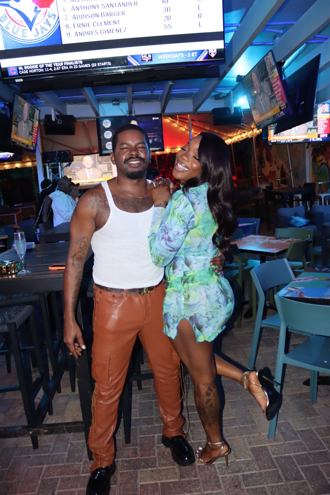 Stylish couple poses together smiling on the Grails patio at night, with MLB Tonight playing on overhead TVs.