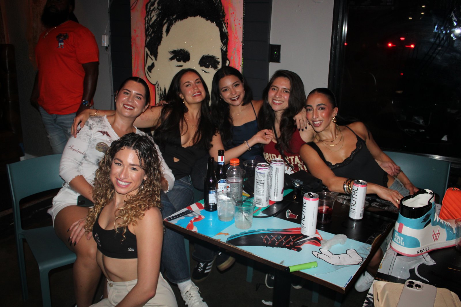 Six women pose together smiling at a Grails Miami table with Michelob Ultra, NUTRL cans and a sneaker-shaped Holy Grail cocktail bucket