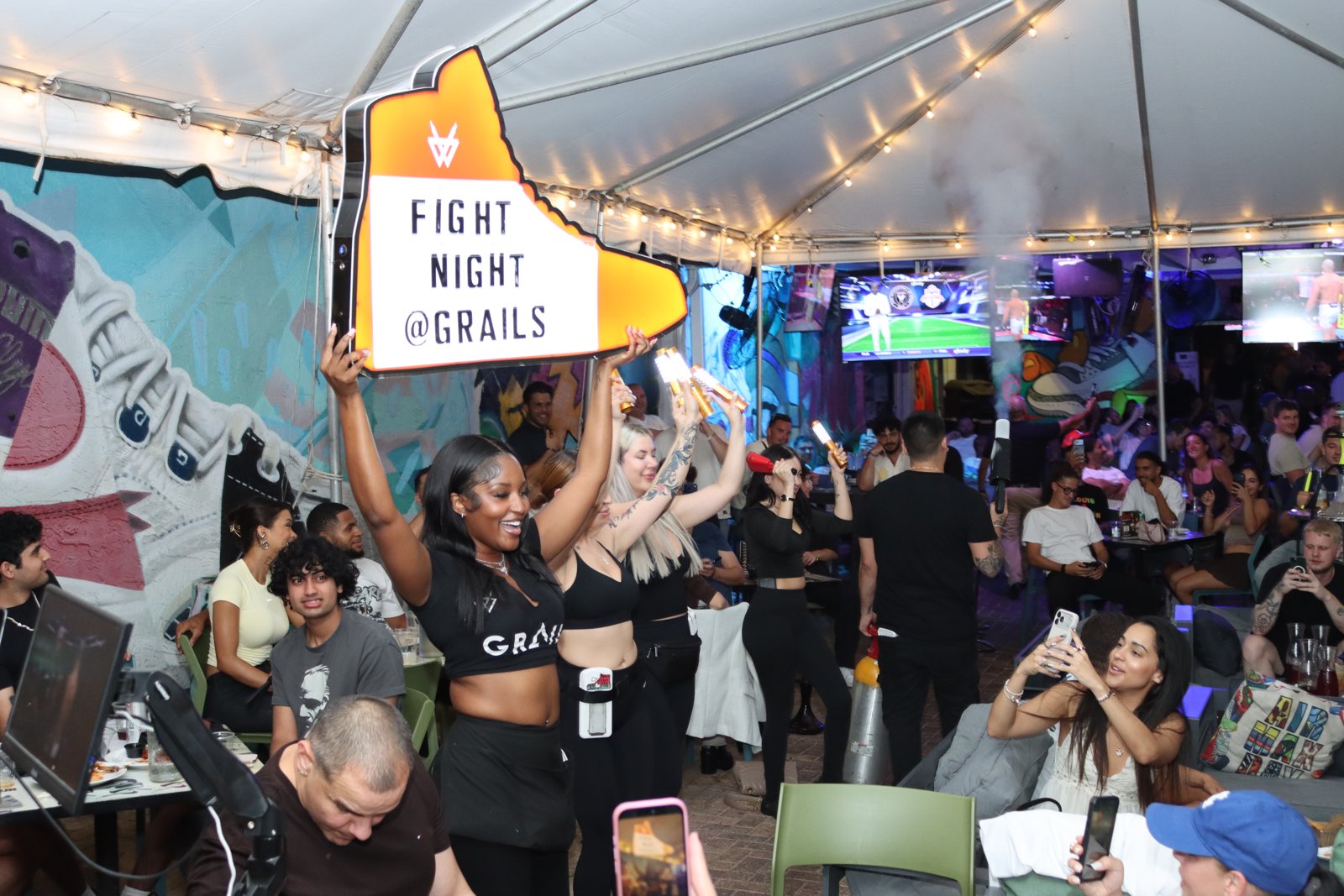 Grails server holds up an illuminated sneaker-shaped 'Fight Night @ Grails' sign while teammates carry sparkler bottles through the patio crowd