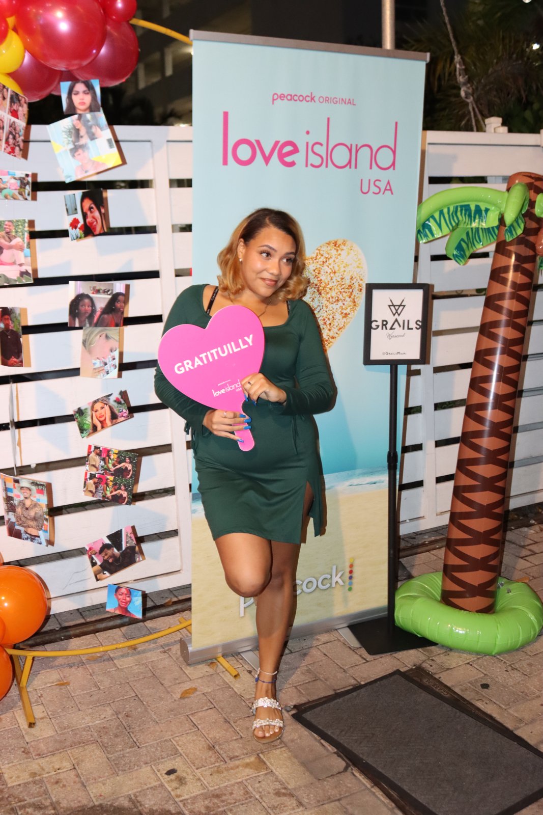 Woman in green dress poses with pink 'Gratituilly' heart paddle at Love Island USA watch party at Grails Miami