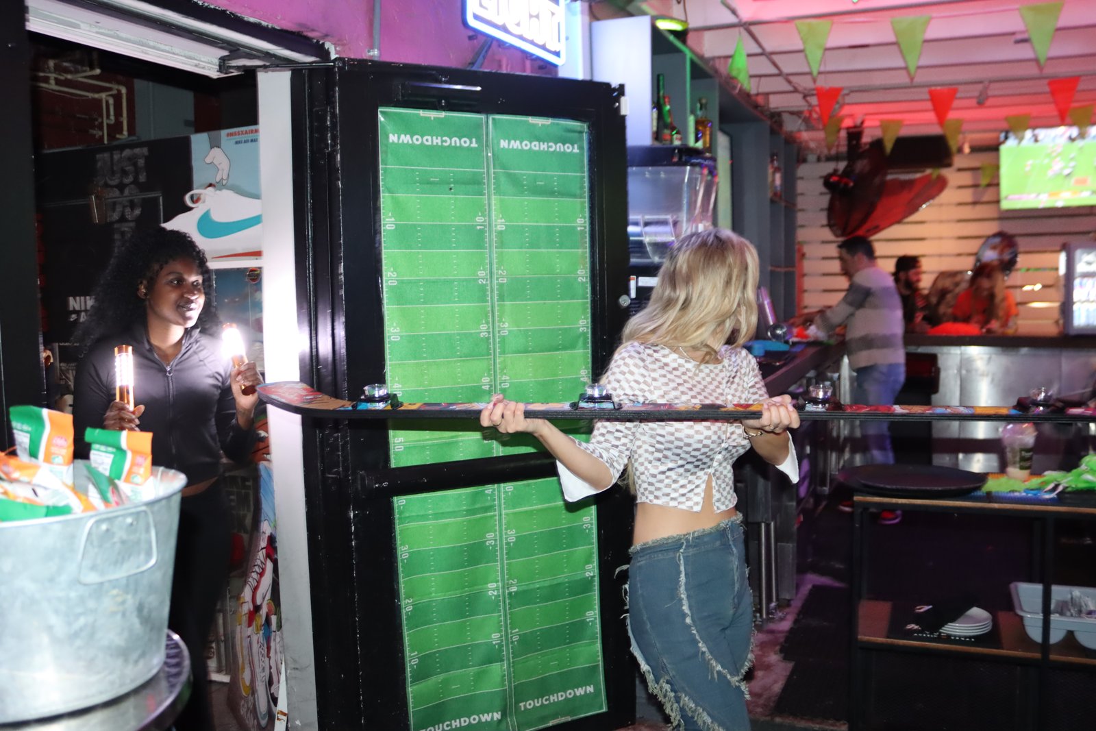 Blonde woman in checkered top holds a shot ski at the bar while a smiling woman with sparkler shots stands beside a football-field-printed door at Grails Miami.
