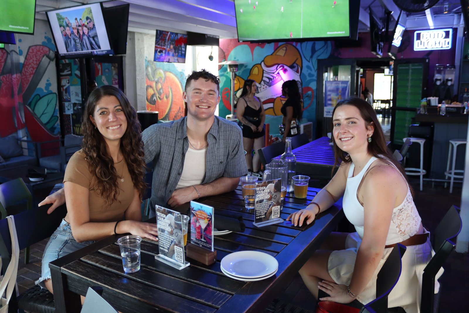 Three friends smile at the camera around a table with beers at Grails Wynwood, sneaker mural and soccer on TV in background