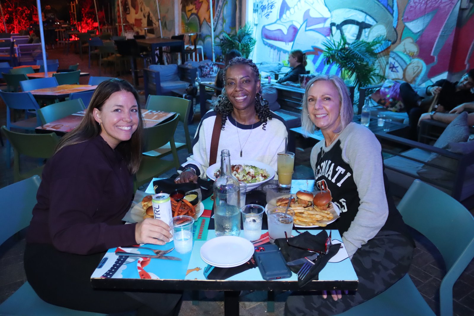 Three women smiling at a patio table at Grails Wynwood with burgers, salad, and fries spread between them