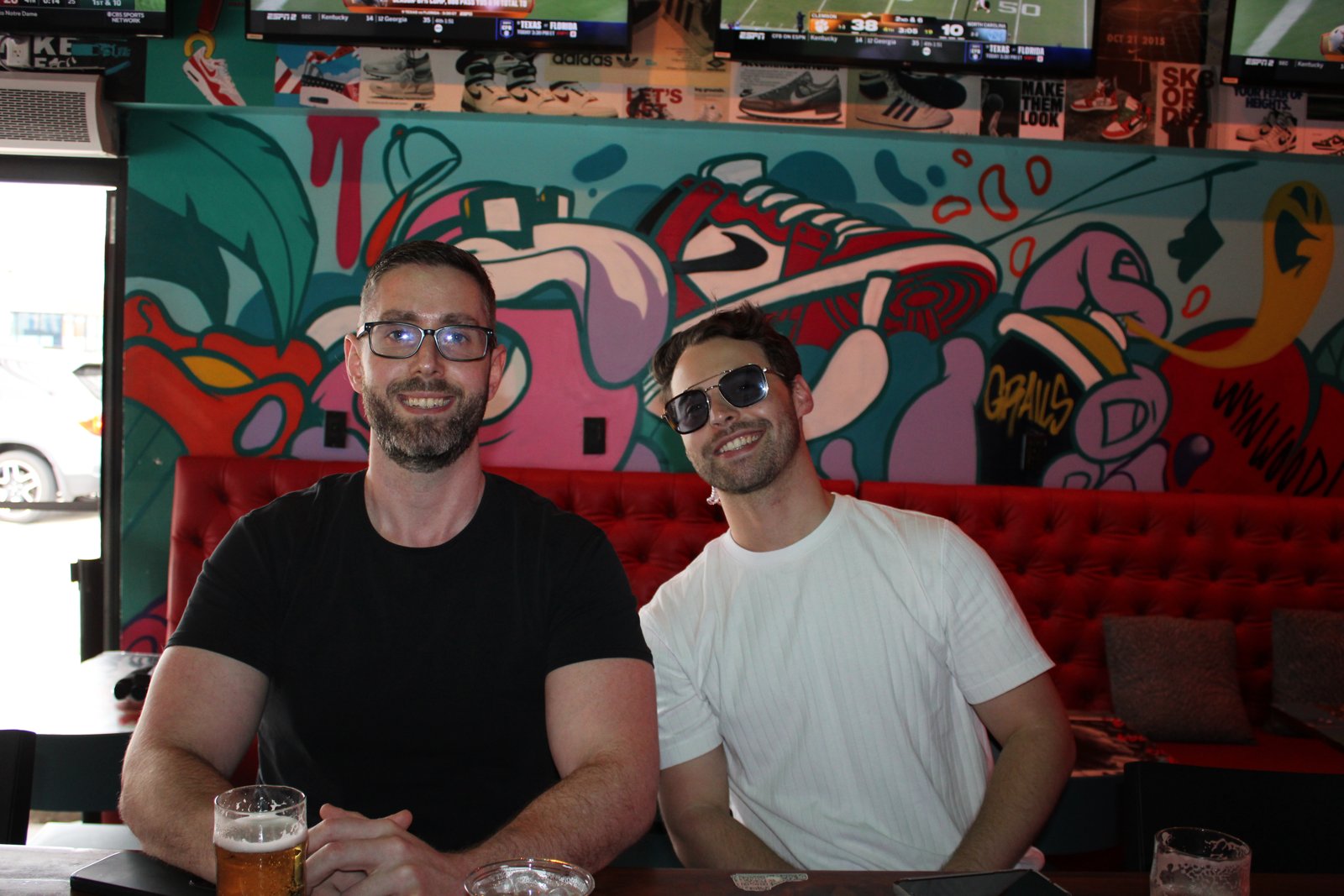 Two smiling men sit at a red booth with beers in front of a colorful Wynwood sneaker mural at Grails.