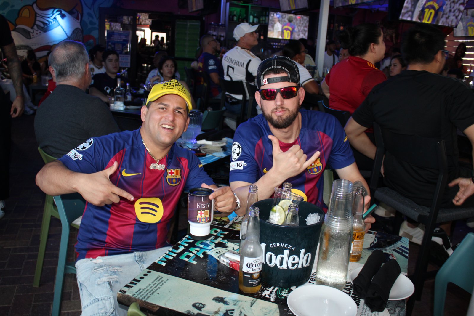 Two FC Barcelona fans in jerseys pose with a Modelo beer bucket at Grails Miami during a soccer watch party.