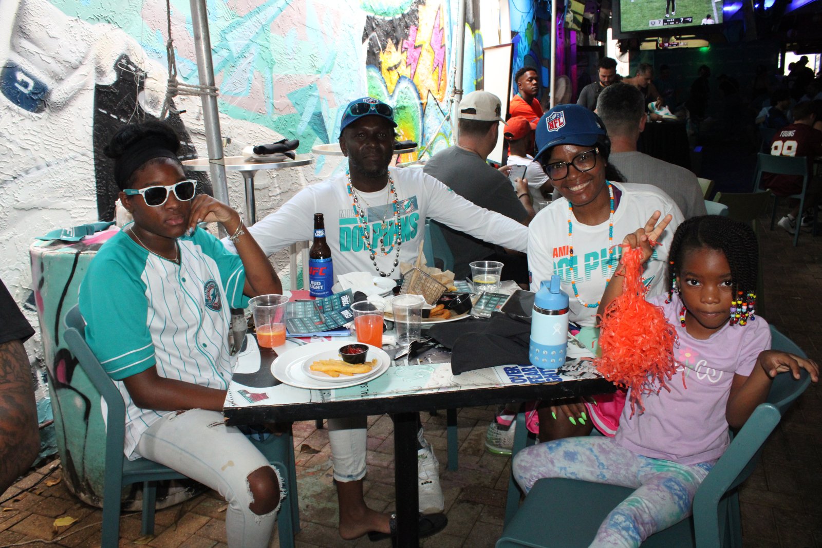 Family in Miami Dolphins gear poses at a Grails patio table with fries, drinks and bingo cards during an NFL watch party.