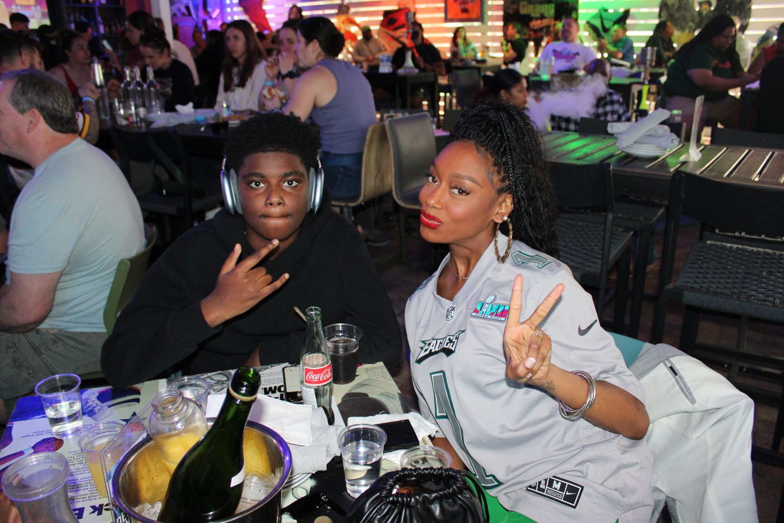 Woman in Philadelphia Eagles Super Bowl LVII jersey poses with peace sign beside young fan in headphones at Grails Miami watch party