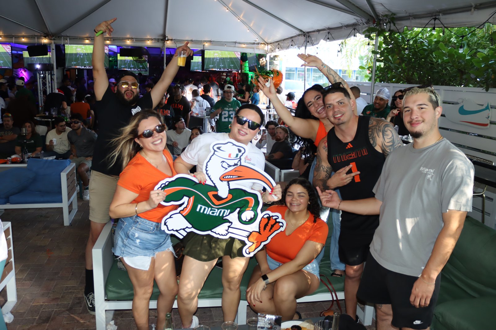Group of Miami Hurricanes fans in orange and green pose with a large Sebastian the Ibis cutout at a Grails watch party on the covered patio.