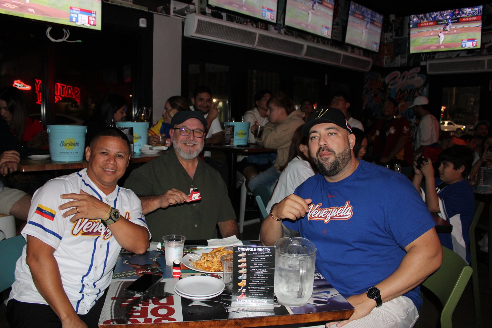 Three Venezuela baseball fans in Vinotinto jerseys and cap pose at a Grails table with a sneaker shot and Sneaker Shots menu during a WBC-style watch party.